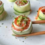 Cucumber and salmon rolls garnished with herbs on a white plate. One roll is being held by wooden chopsticks in the foreground.
