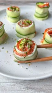 Cucumber and salmon rolls garnished with herbs on a white plate. One roll is being held by wooden chopsticks in the foreground.