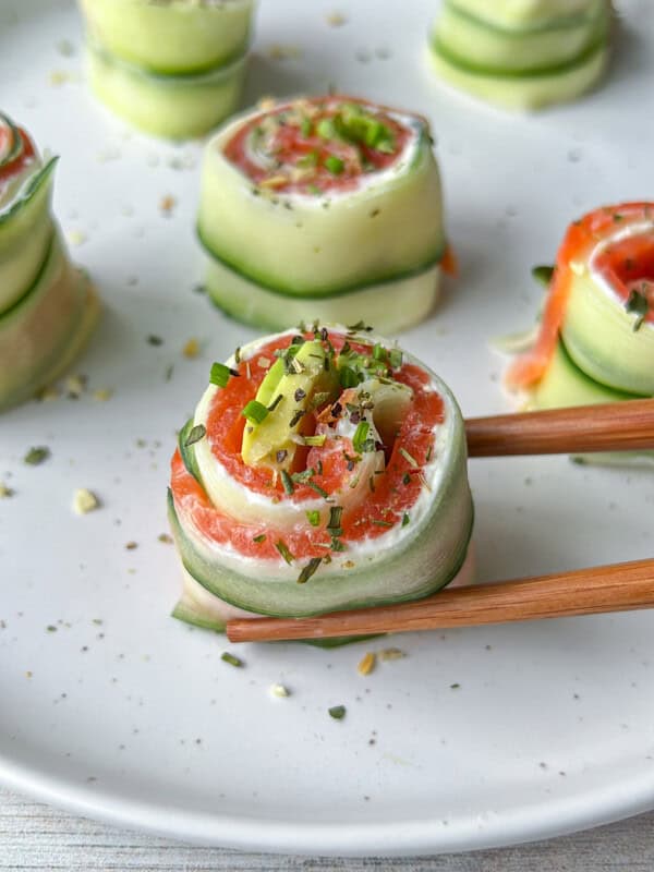 Cucumber and salmon rolls garnished with herbs on a white plate. One roll is being held by wooden chopsticks in the foreground.
