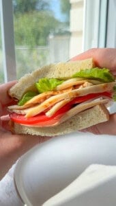 A close-up of a hand holding a sandwich with sliced turkey, tomato, lettuce, and cheese between two thick slices of bread, with a blurred window in the background.