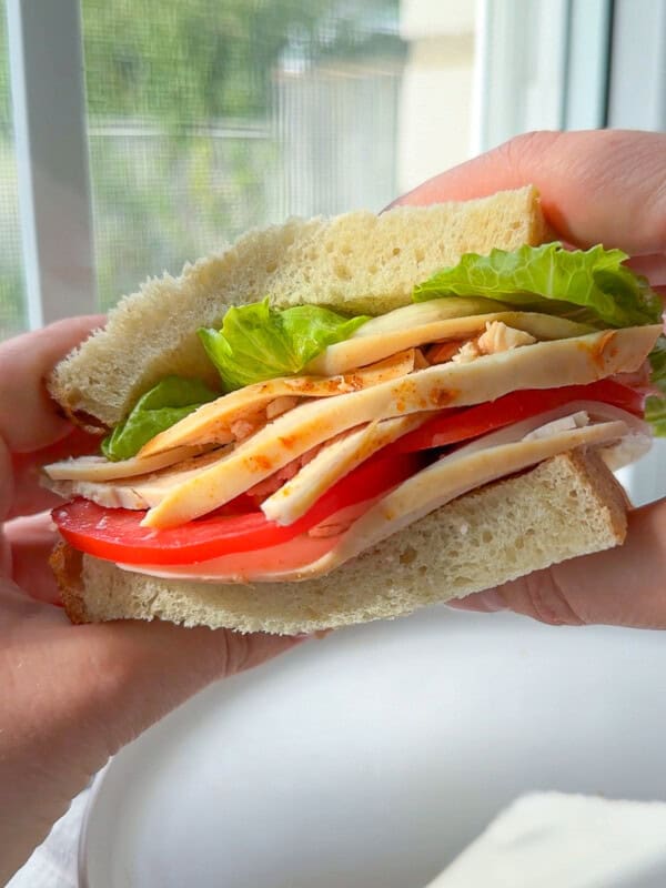 A close-up of a hand holding a sandwich with sliced turkey, tomato, lettuce, and cheese between two thick slices of bread, with a blurred window in the background.