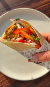 A hand holding a pita wrap filled with falafel, sliced tomatoes, onions, and fresh herbs, wrapped in paper with text, with a white plate and wooden table in the background.