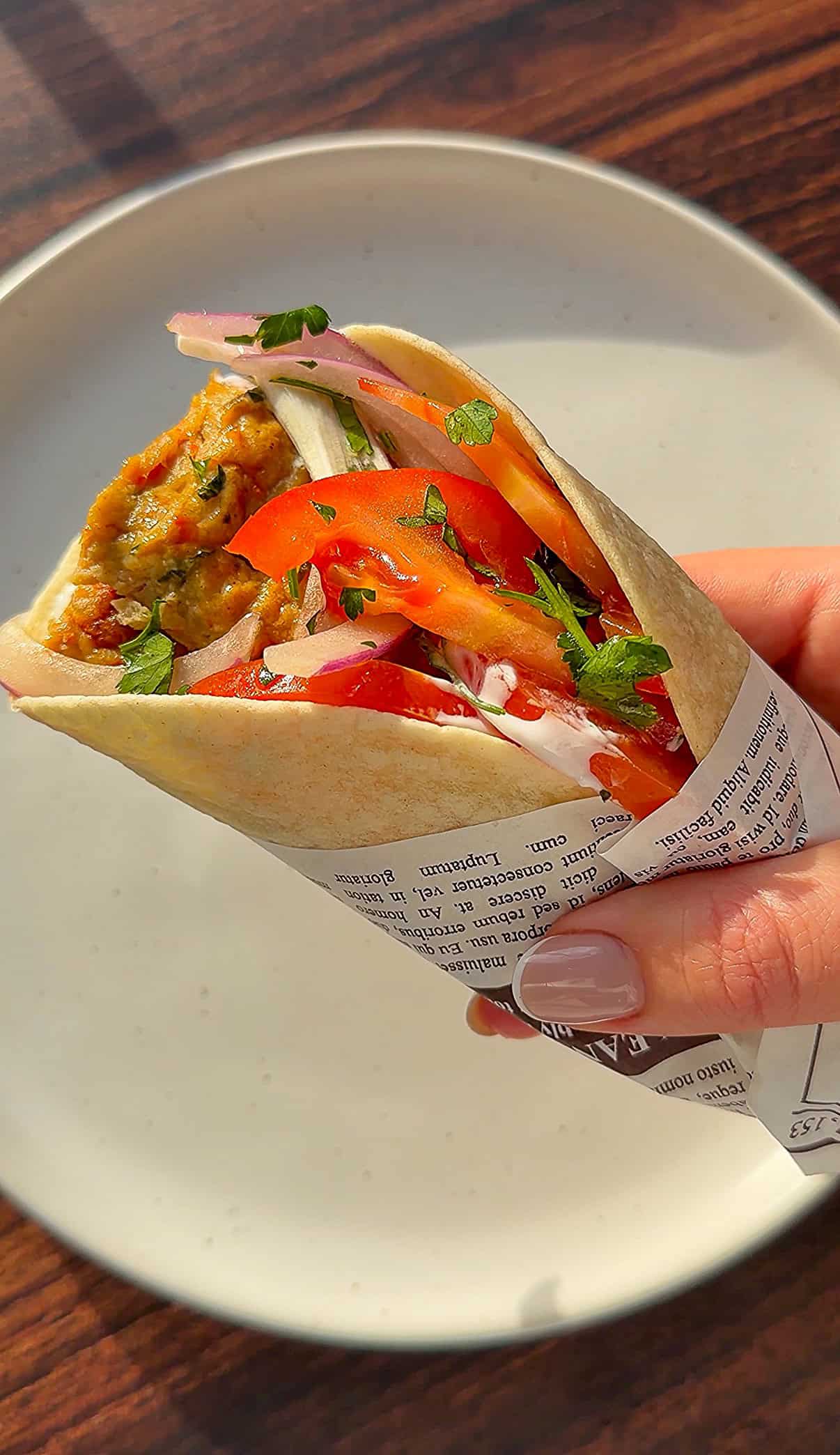 A hand holding a pita wrap filled with falafel, sliced tomatoes, onions, and fresh herbs, wrapped in paper with text, with a white plate and wooden table in the background. - 4