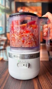 A hand holds the lid of a KitchenAid food processor filled with chopped onions, tomatoes, and other vegetables, set on a wooden table with a blurred café background.