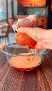 A hand squeezes a mesh bag filled with blended tomatoes over a glass bowl, straining out tomato juice onto a wooden surface.