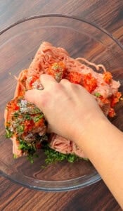 A hand mixes ground meat, chopped herbs, and spices in a clear glass bowl on a wooden surface.