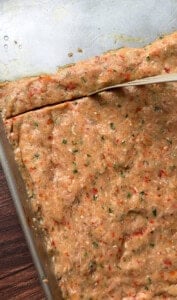 A pan filled with a thick, uncooked, herbed bread or dough mixture, speckled with red and green seasoning, being sliced with a knife on a wooden surface.