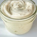 A small glass jar filled with creamy, swirled white sauce or dip, possibly mayonnaise or aioli, set on a white surface with bright, natural lighting.