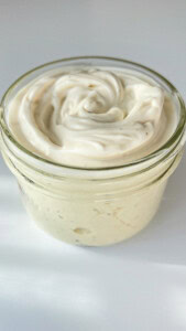 A small glass jar filled with creamy, swirled white sauce or dip, possibly mayonnaise or aioli, set on a white surface with bright, natural lighting.