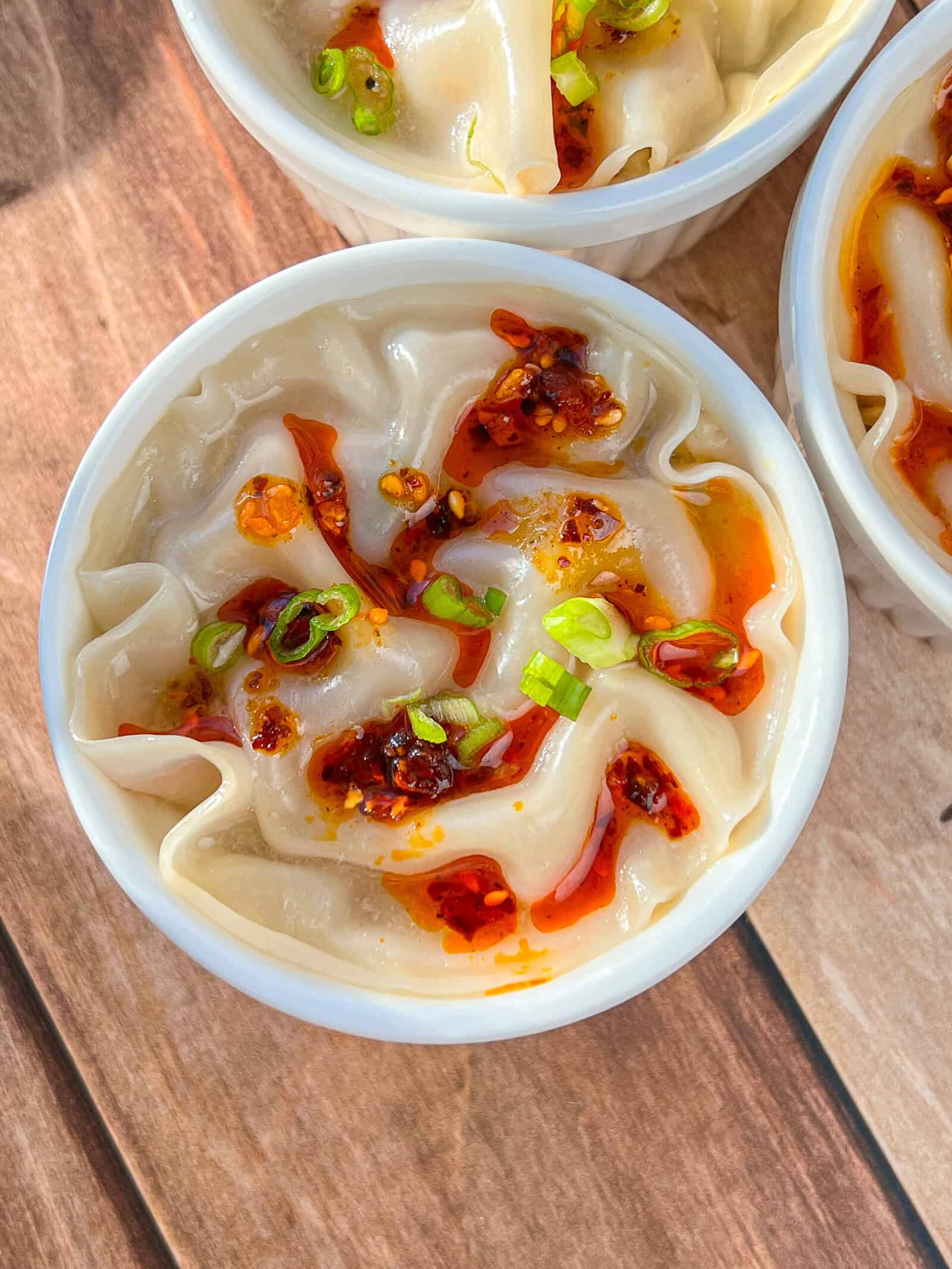 A close-up of a white bowl filled with dumplings topped with chili oil and sliced green onions, placed on a wooden surface. - 3