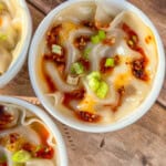 A bowl of dumplings topped with chili oil and chopped green onions, placed on a wooden surface.