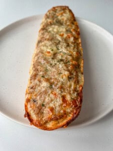 A close-up of a baked, cheese-covered piece of garlic bread on a white plate, with melted, slightly browned cheese and herbs sprinkled on top.