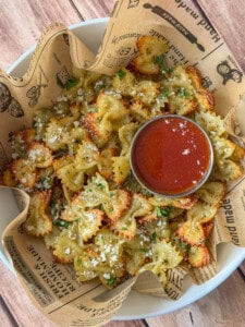 A bowl lined with printed parchment holds crispy bowtie pasta chips sprinkled with grated cheese and herbs, served with a small dish of red marinara sauce for dipping.