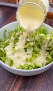A bowl of shredded lettuce on a wooden table, with creamy dressing being poured over it from a glass container.