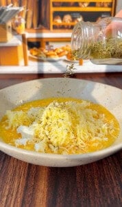 A hand sprinkles dried herbs from a jar onto a bowl with beaten eggs and shredded cheese, set on a wooden table with a bakery counter blurred in the background.