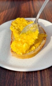 A spoon spreads creamy scrambled eggs onto a slice of bread on a white plate atop a wooden table.