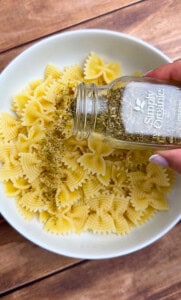 A hand pours dried oregano from a Simply Organic jar onto a bowl of cooked farfalle pasta on a wooden surface.
