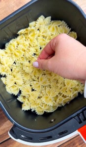 A hand sprinkles seasoning onto uncooked bow-tie pasta in an air fryer basket. The pasta is arranged in a single layer, ready to be cooked.