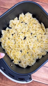 Uncooked farfalle pasta seasoned with herbs and spices is spread evenly in the basket of an air fryer, ready for cooking. A hand is holding the edge of the basket, and the background is a wooden surface.