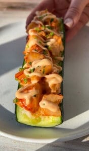 A hand holds a halved zucchini filled with diced vegetables, topped with a creamy sauce and chopped herbs, on a white plate in natural light.