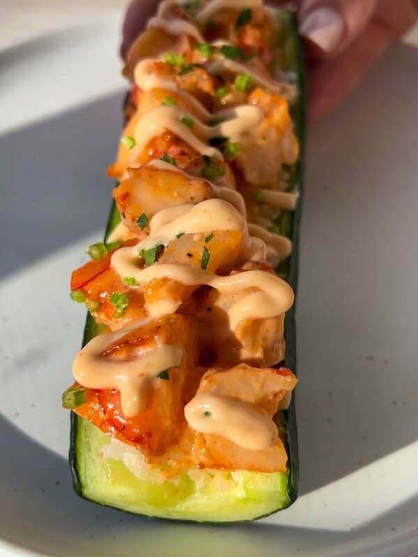 A hand holds a halved zucchini filled with diced vegetables, topped with a creamy sauce and chopped herbs, on a white plate in natural light. - 9