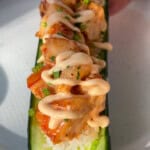 A hand holds a halved cucumber filled with sushi rice, topped with grilled shrimp, chopped herbs, and a drizzle of creamy sauce, served on a white plate in bright sunlight.