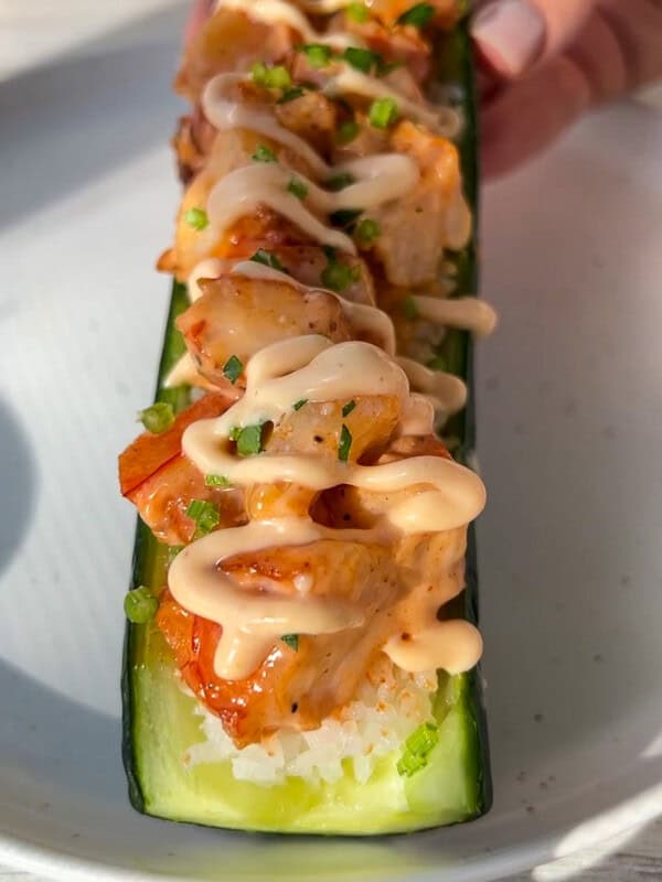 A hand holds a halved cucumber filled with sushi rice, topped with grilled shrimp, chopped herbs, and a drizzle of creamy sauce, served on a white plate in bright sunlight.