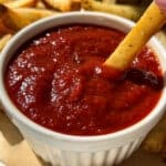 A hand dips a French fry into a small white ramekin filled with red ketchup, with more fries on a plate in the background.