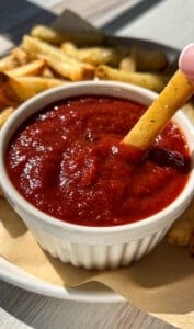A hand dips a French fry into a small white ramekin filled with red ketchup, with more fries on a plate in the background.