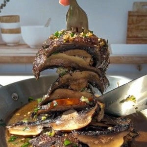 A hand holding a fork lifts a stack of sautéed portobello mushroom caps covered in herbs and sauce from a skillet, with a spoon resting in the pan and a bright kitchen background.