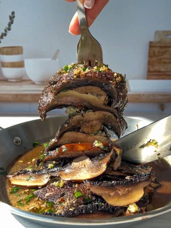 A hand holding a fork lifts a stack of sautéed portobello mushroom caps covered in herbs and sauce from a skillet, with a spoon resting in the pan and a bright kitchen background.