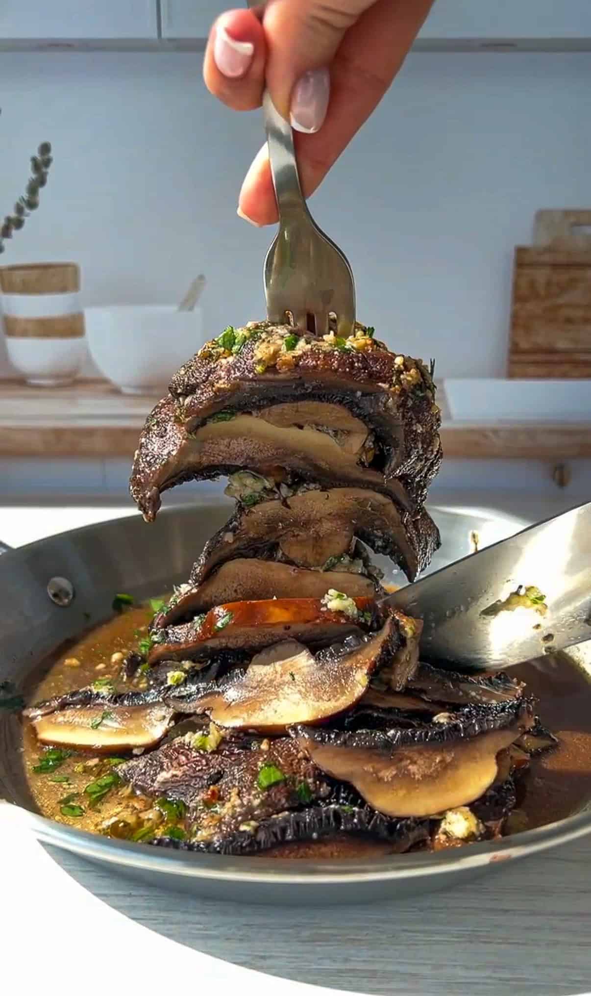 A hand holding a fork lifts a stack of sautéed portobello mushroom caps covered in herbs and sauce from a skillet, with a spoon resting in the pan and a bright kitchen background. - 3