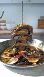 A stack of grilled portobello mushroom slices, skewered vertically in a pan with rich sauce, sits on a kitchen counter. Sunlight highlights the mushrooms and sauce, with bowls and wooden decor in the background.