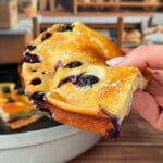 A hand holds up a slice of fluffy blueberry cake dusted with powdered sugar, with a pan of more cake and shelves of bread in the blurred background.