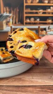 A hand holds up a slice of fluffy blueberry cake dusted with powdered sugar, with a pan of more cake and shelves of bread in the blurred background.
