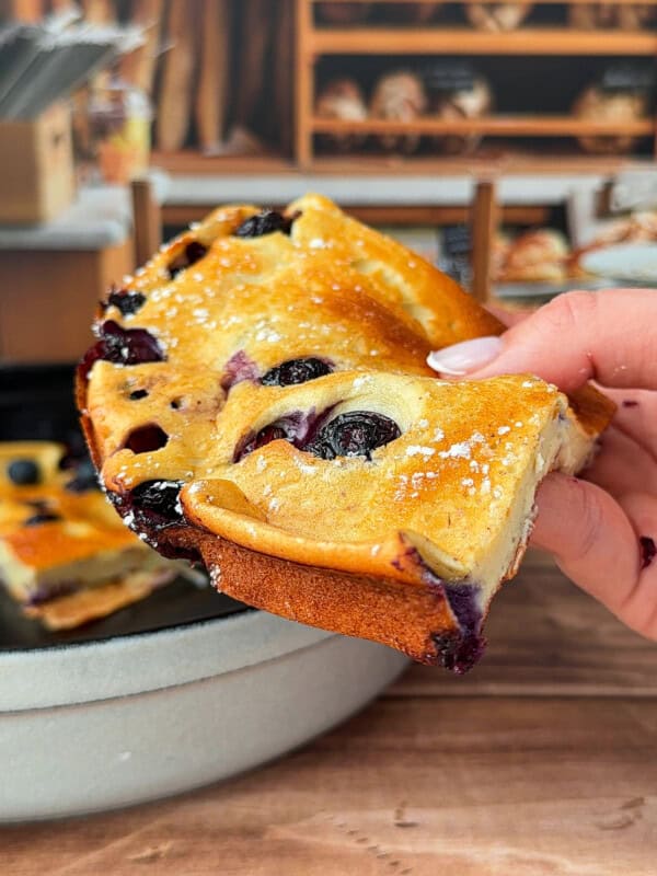 A hand holds up a slice of fluffy blueberry cake dusted with powdered sugar, with a pan of more cake and shelves of bread in the blurred background.