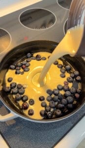 Batter is being poured into a cast iron skillet filled with fresh blueberries, sitting on a stove with a glass cooktop.
