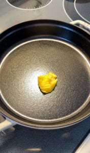 A small dollop of yellow butter sits in the center of a black cast iron skillet on a stovetop, ready to be melted before cooking.