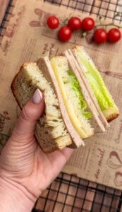 A hand holds a sandwich with thick bread slices, layers of ham, cheese, and lettuce. In the background, a few cherry tomatoes rest on parchment paper.