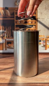A hand holds a spiral whisk above a tall, stainless steel container on a wooden surface, with a bakery display of breads and pastries blurred in the background.