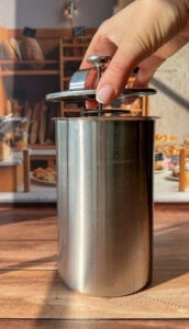 A hand presses down the plunger of a stainless steel French press on a wooden surface, with a bakery display blurred in the background.