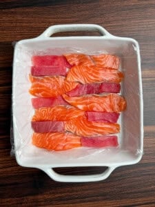 A white square dish lined with plastic wrap holds alternating rows of sliced raw salmon and tuna, displayed neatly on a dark wooden surface.