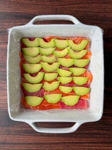 A square white dish lined with plastic wrap, filled with overlapping slices of raw salmon, raw tuna, and avocado arranged in neat rows on a dark wood surface.