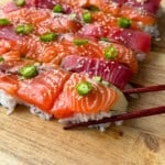 Rectangular pieces of sushi with salmon and tuna slices on rice, topped with green chili slices and sesame seeds, arranged on a wooden board. A pair of chopsticks holds one piece.