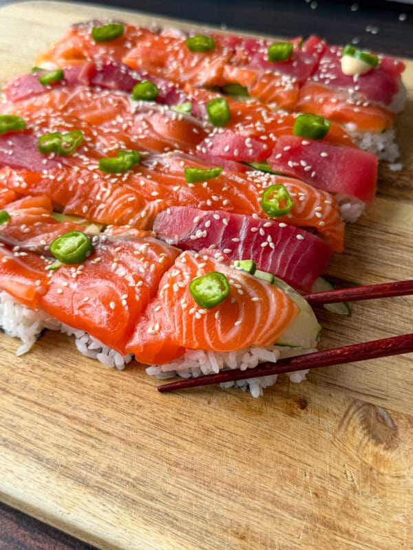 Rectangular pieces of sushi with salmon and tuna slices on rice, topped with green chili slices and sesame seeds, arranged on a wooden board. A pair of chopsticks holds one piece.