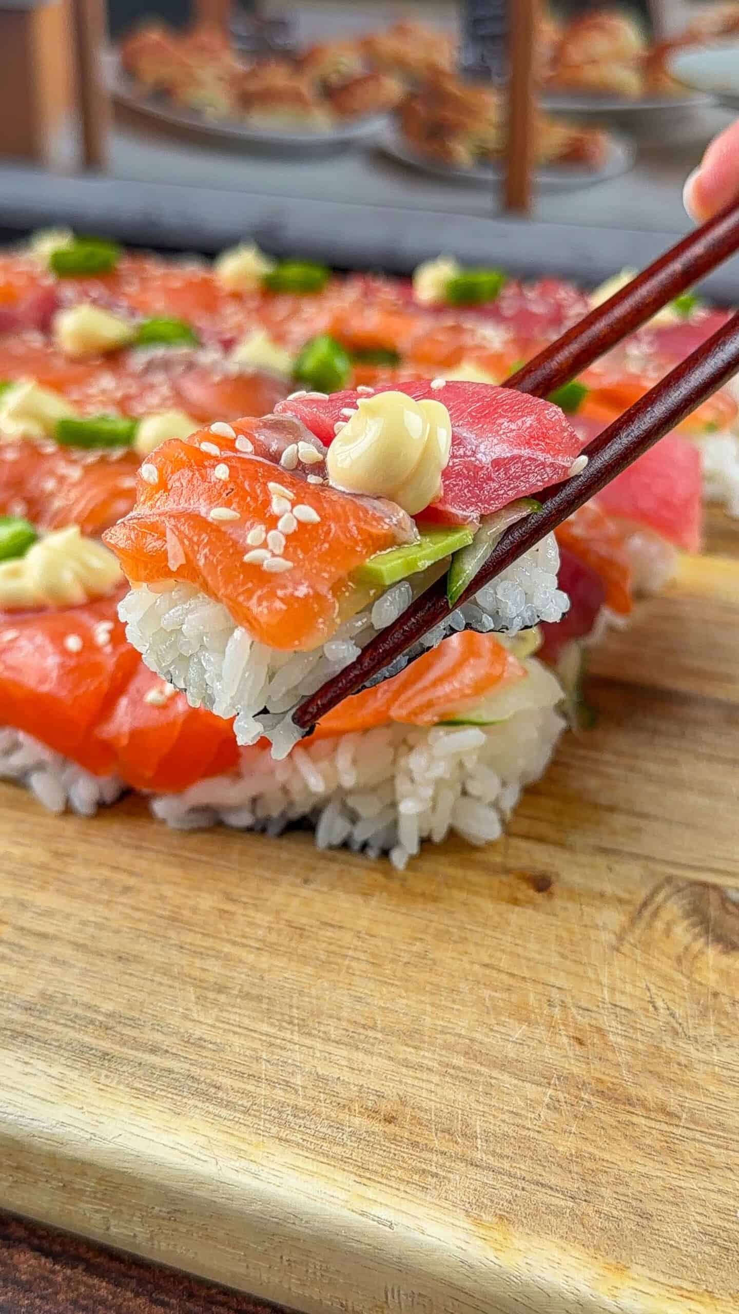 A close-up of chopsticks holding a piece of sushi with rice, fresh salmon, tuna, green onions, sesame seeds, and a dollop of mayonnaise, above a wooden board with more sushi in the background.