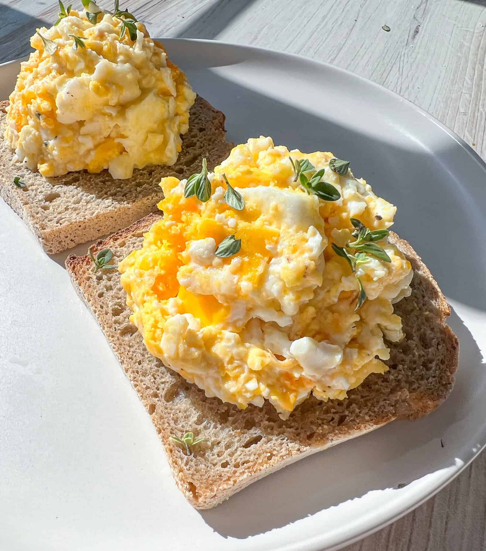 Two slices of whole grain bread topped with scrambled eggs and garnished with fresh herbs, served on a white plate in bright natural light. - 4