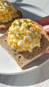 A hand holds a slice of bread topped with a generous scoop of egg salad, garnished with small green herb sprigs, on a white plate in bright natural light.