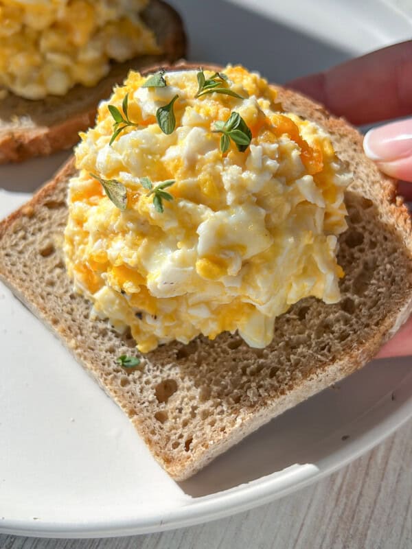 A hand holds a slice of bread topped with a generous scoop of egg salad, garnished with small green herb sprigs, on a white plate in bright natural light.