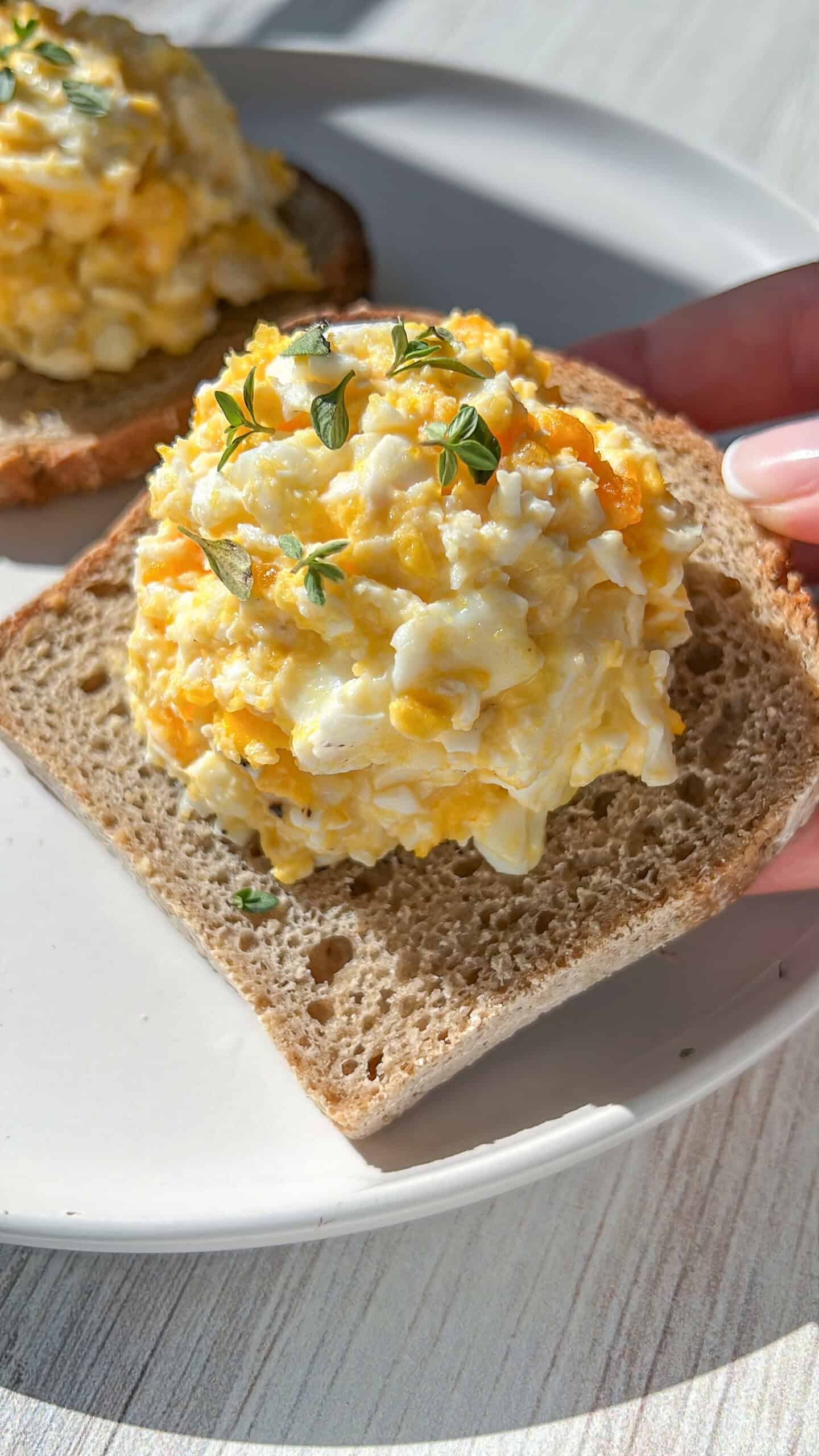 A hand holds a slice of bread topped with a generous scoop of egg salad, garnished with small green herb sprigs, on a white plate in bright natural light. - 3